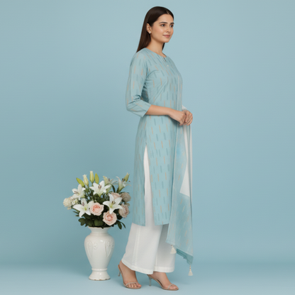 Woman in a light blue and white traditional outfit standing next to a wooden chair and table with a floral arrangement on a blue background.