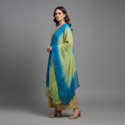 A woman modeling a traditional Indian outfit consisting of a light olive green kurta with gold stripes, matching brown pants, and a blue-green gradient dupatta, set against a solid gray studio background.