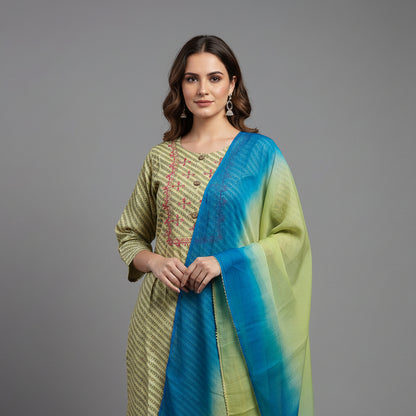A woman modeling a traditional Indian outfit consisting of a light olive green kurta with gold stripes, matching brown pants, and a blue-green gradient dupatta, set against a solid gray studio background.