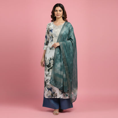 A woman modeling a traditional Indian outfit consisting of a long-sleeved kurta with a white, gray, and blue floral print, a matching teal-green shawl draped over her shoulders, and navy blue pants, set against a solid pink studio background.