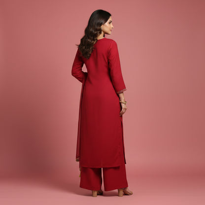 A woman modeling a traditional red Indian salwar kameez suit with intricate gold, green, and blue embroidery on the chest and border, paired with a matching dupatta, against a neutral gray studio background.