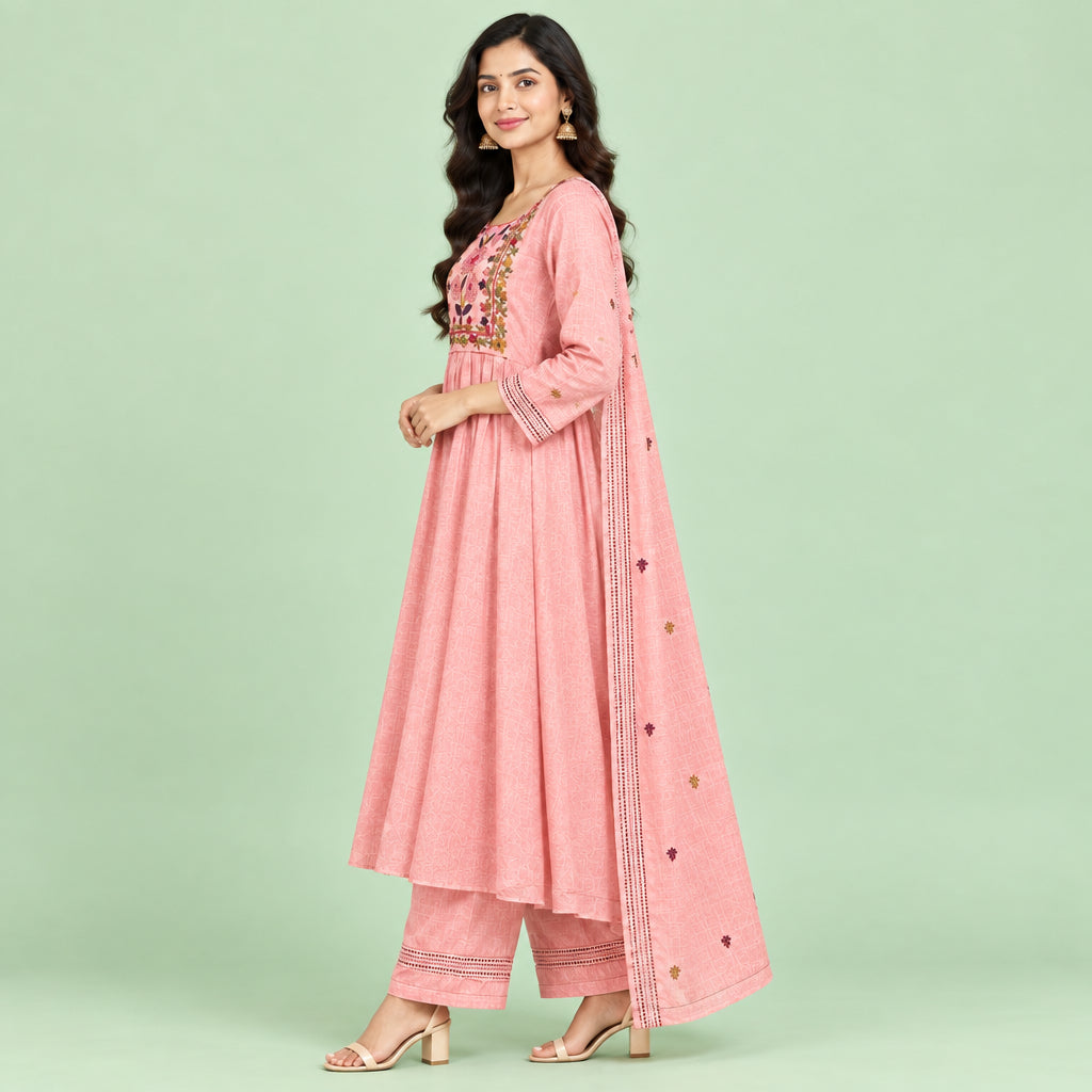 A pink traditional women's outfit laid flat on a patterned bedspread, consisting of a long kurta with floral embroidery around the neckline, matching trousers, and a sheer skirt or drape piece with small embroidered details.