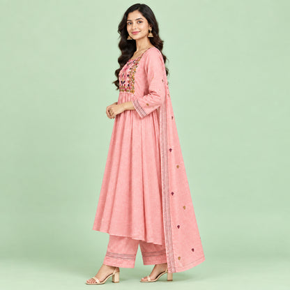 A pink traditional women's outfit laid flat on a patterned bedspread, consisting of a long kurta with floral embroidery around the neckline, matching trousers, and a sheer skirt or drape piece with small embroidered details.