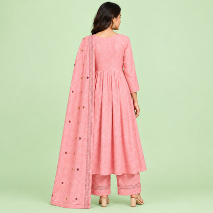 A pink traditional women's outfit laid flat on a patterned bedspread, consisting of a long kurta with floral embroidery around the neckline, matching trousers, and a sheer skirt or drape piece with small embroidered details.