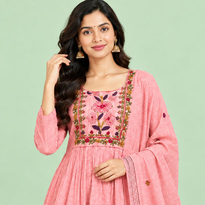 A pink traditional women's outfit laid flat on a patterned bedspread, consisting of a long kurta with floral embroidery around the neckline, matching trousers, and a sheer skirt or drape piece with small embroidered details.