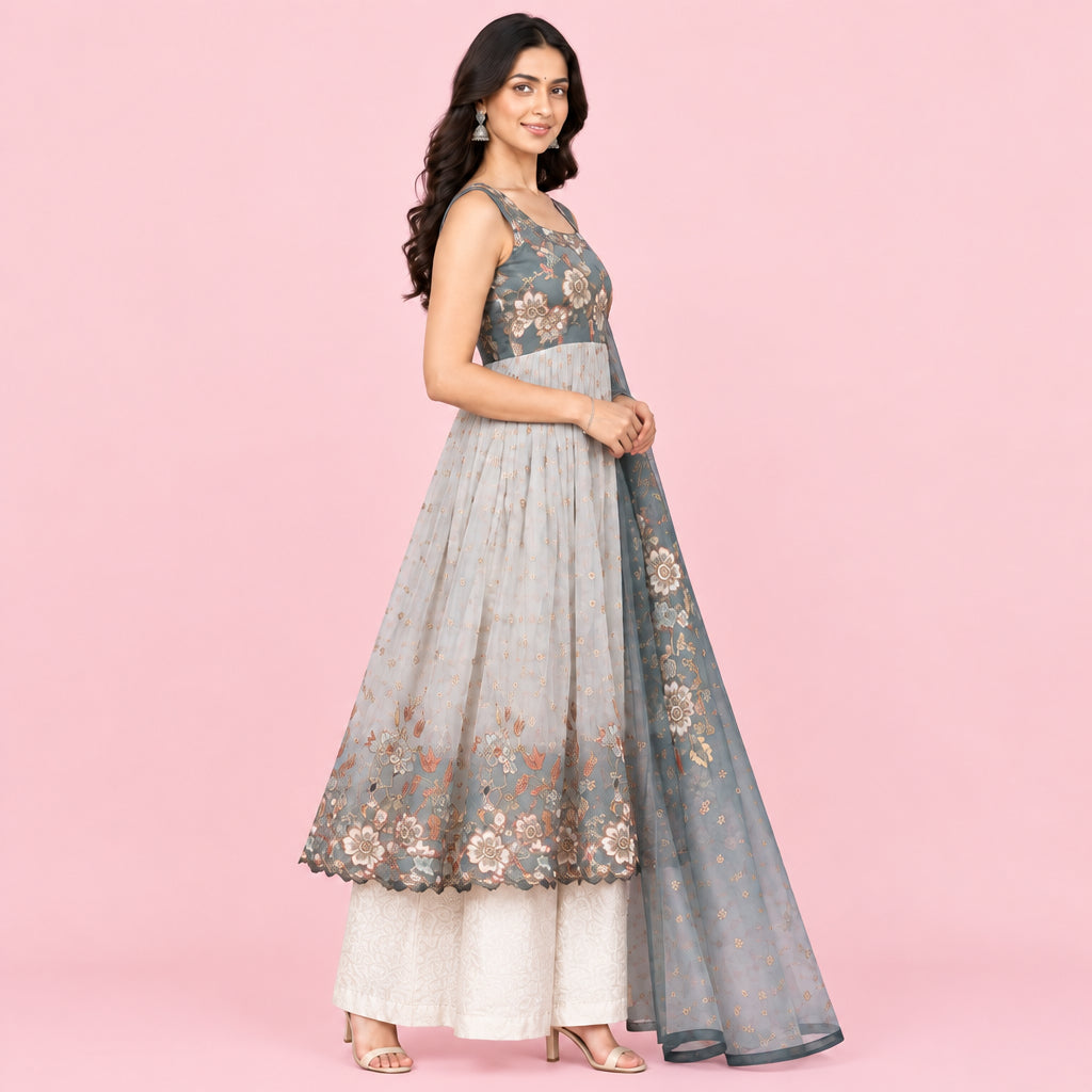 A woman models a traditional Indian outfit consisting of a light gray-blue anarkali kurta with floral embroidery, a matching dupatta, and cream-colored palazzo pants, set against an elegant courtyard background.