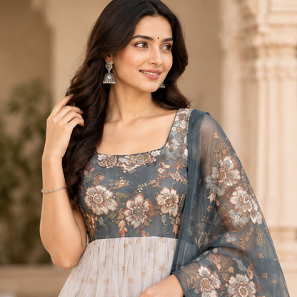 A woman models a traditional Indian outfit consisting of a light gray-blue anarkali kurta with floral embroidery, a matching dupatta, and cream-colored palazzo pants, set against an elegant courtyard background.