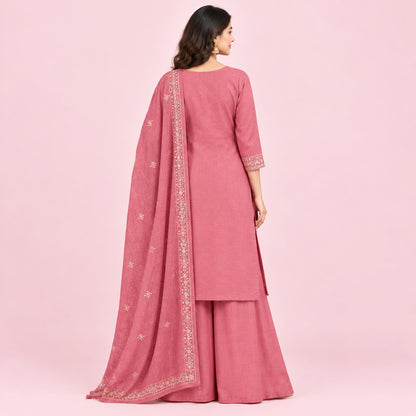 A woman models a dusty pink Indian ethnic suit set featuring a kurta with gold embroidery, matching wide-leg pants, and a sheer dupatta with gold border detailing, set against a neutral beige background.
