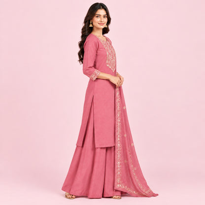A woman models a dusty pink Indian ethnic suit set featuring a kurta with gold embroidery, matching wide-leg pants, and a sheer dupatta with gold border detailing, set against a neutral beige background.