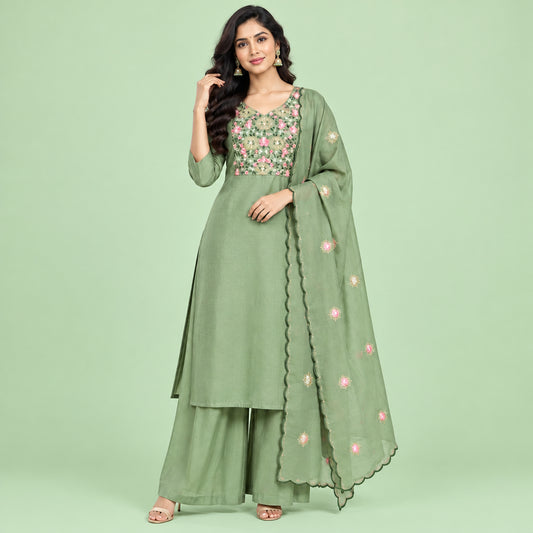 A sage green three-piece traditional outfit set consisting of a long kurta with floral embroidery on the chest, matching flared pants, and a folded skirt, laid out on a bed.