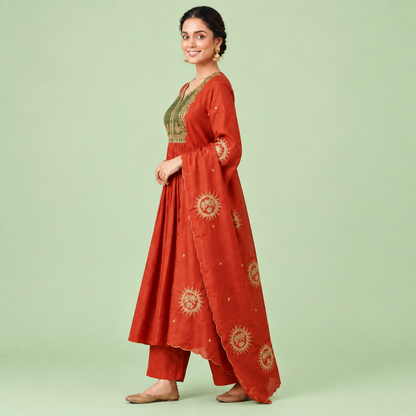 A woman models a rust-orange traditional Indian outfit consisting of a long-sleeved kurta with an olive green embroidered yoke, matching pleated palazzo pants, and a coordinating dupatta decorated with gold sun motifs, set against a solid light green background.