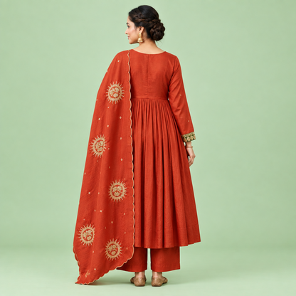 A woman models a rust-orange traditional Indian outfit consisting of a long-sleeved kurta with an olive green embroidered yoke, matching pleated palazzo pants, and a coordinating dupatta decorated with gold sun motifs, set against a solid light green background.